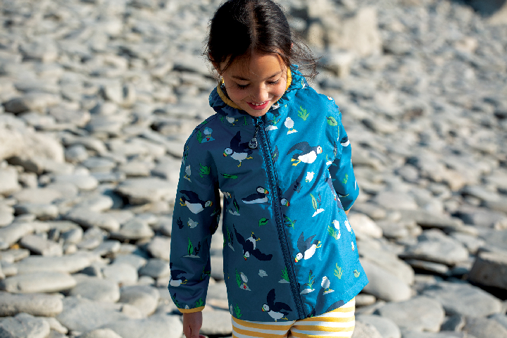 Yound Dark Girl with dark hair with a blue Pioneer Packaway Jacket, with picturs of puffins on it, as she walks on a pebble beach.